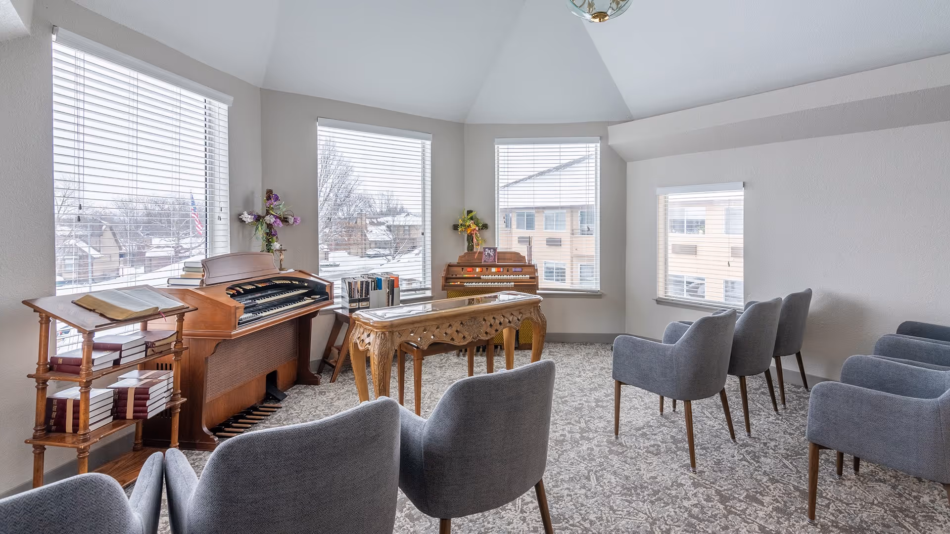 A bright room with large windows featuring white blinds, containing two wooden organs, a wooden table with intricate carvings, a small bookshelf with books, and several gray upholstered chairs arranged in rows facing the organs.