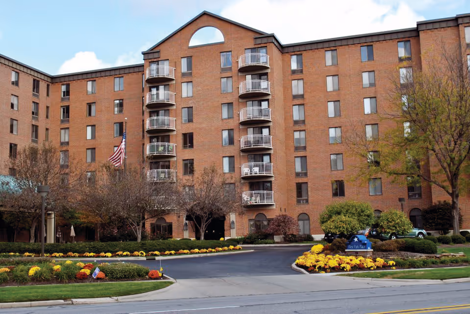 Exterior view of a multi-story brick senior living facility with balconies, landscaped flower beds, trees, and an American flag near the entrance driveway.