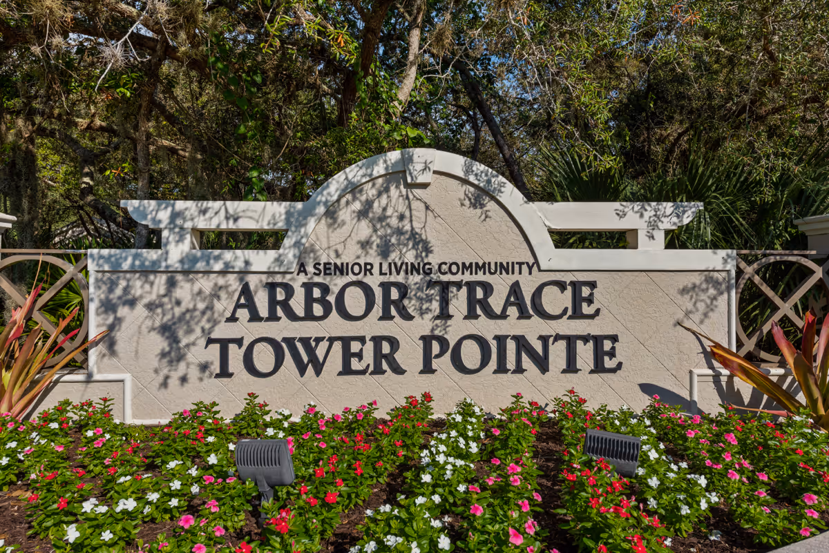Entrance sign for Arbor Trace Tower Pointe, a senior living community, surrounded by colorful flowers and greenery with trees in the background.