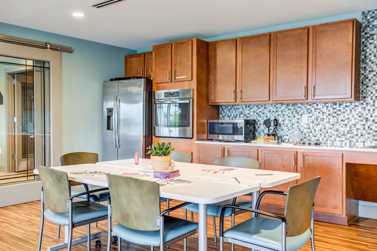 A bright kitchen area with wooden cabinets, a stainless steel refrigerator, built-in oven, microwave, and a mosaic tile backsplash. In front of the kitchen is a white table with six chairs around it, some art supplies and a small potted plant on the table. The floor is wooden and there is a glass door on the left side.