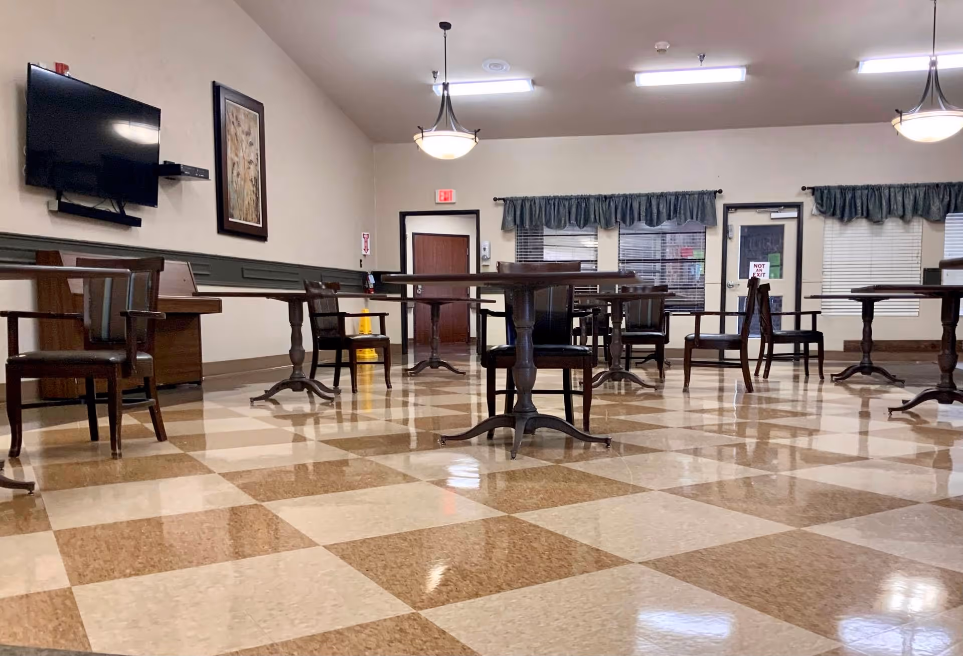 A spacious dining room with checkered brown and beige tile flooring, several round wooden tables with chairs arranged around them, a wall-mounted flat screen TV, framed artwork, and windows with green valances. The room is well-lit with ceiling lights and has an exit door with a 'Not an Exit' sign.