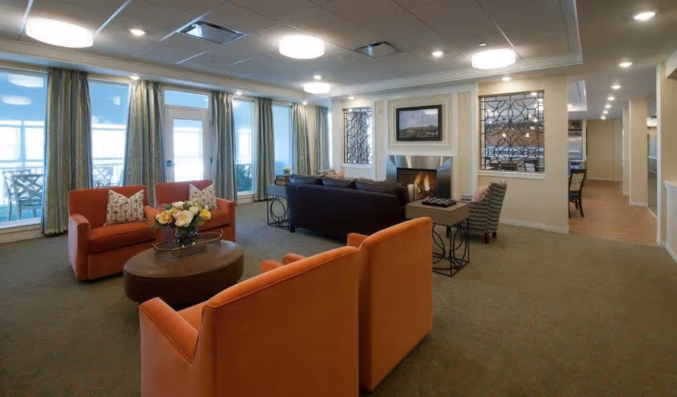 A spacious living room area with orange armchairs and a dark sofa arranged around a round coffee table with a flower arrangement. Large windows with curtains let in natural light, and a fireplace with a framed picture above it is visible. The room has a carpeted floor and recessed ceiling lights.