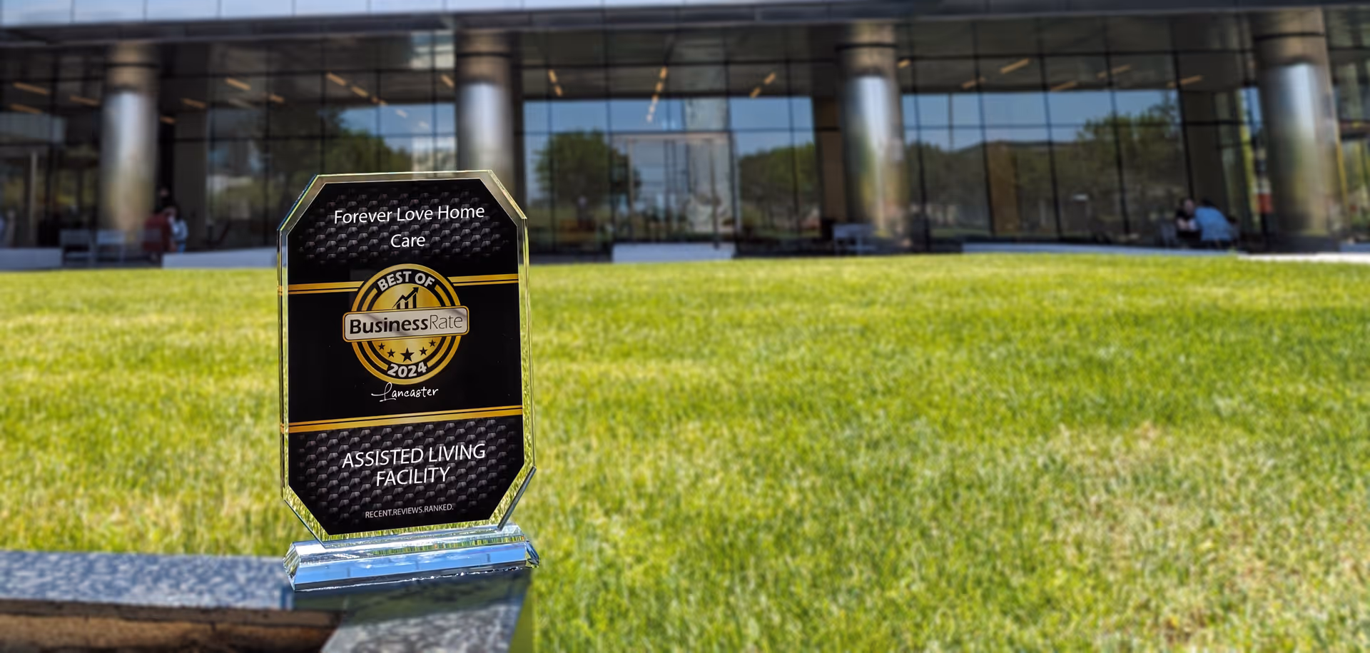A clear award plaque for Forever Love Home Care, Inc. displayed outdoors on a stone ledge with a green lawn and a modern glass building in the background. The plaque reads 'Best of Business Rate 2024 Lancaster Assisted Living Facility'.