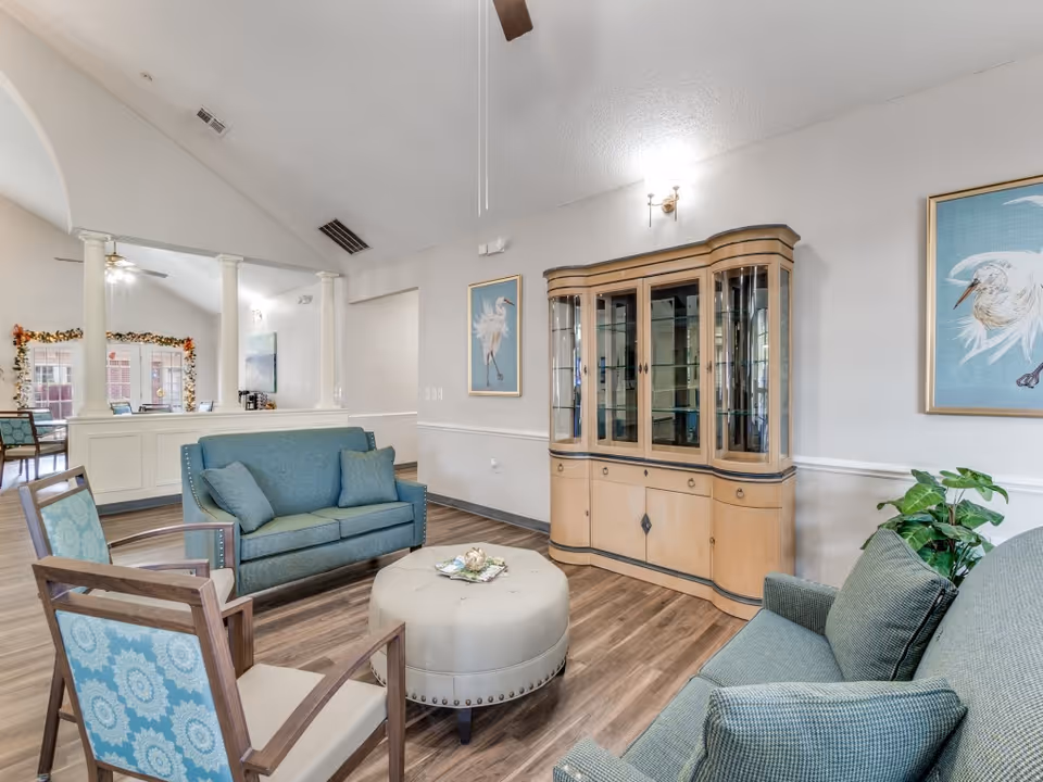 A bright and spacious living room area in a senior living facility with light wood flooring, two teal upholstered sofas, two wooden armchairs with blue patterned cushions, and a round beige ottoman in the center. A wooden display cabinet with glass doors is against the wall, flanked by two framed bird paintings. The room has white walls, a ceiling fan, and an open view into an adjacent room with large windows and garland decorations.