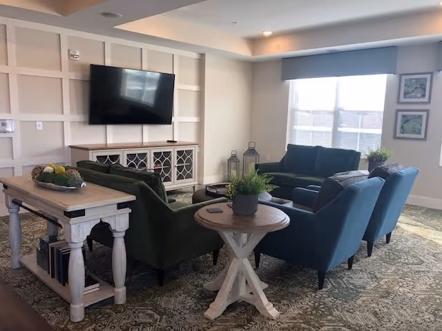 A cozy living room area in a senior living facility featuring a wall-mounted flat screen TV above a white cabinet. The room has two green sofas and two blue armchairs arranged around a central coffee table. There is a wooden console table with books underneath and decorative items on top. A round side table with a potted plant is also visible. Large windows with blinds allow natural light to fill the room, and the floor is covered with a patterned carpet.