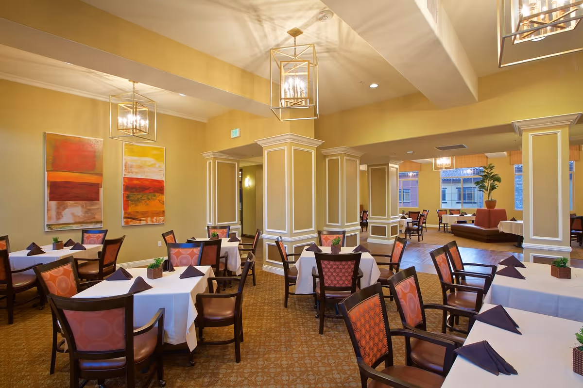 A spacious dining room with multiple tables covered in white tablecloths and set with dark folded napkins. The chairs have wooden frames with patterned upholstery. The room features beige walls with white trim, decorative columns, modern pendant lighting, and colorful abstract paintings on the wall. Large windows let in natural light, and there are potted plants adding greenery to the space.