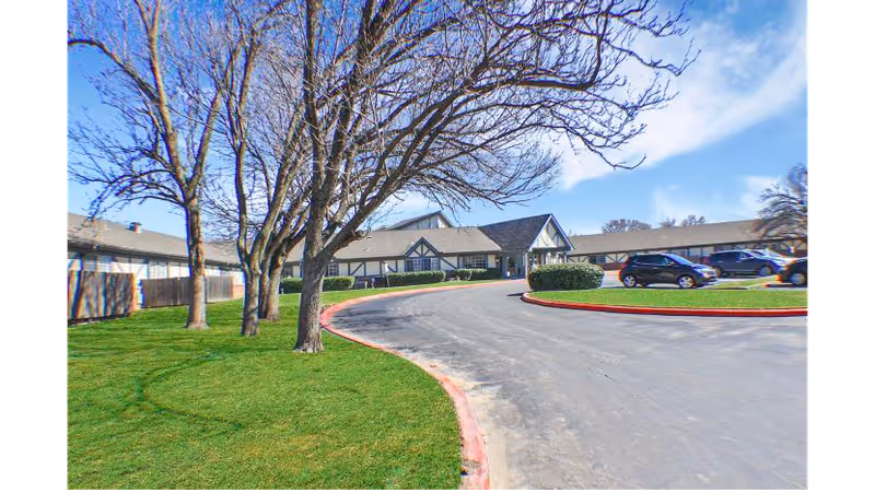 Exterior view of The Cottage At Woodland Hills facility showing a curved driveway, green lawn, leafless trees, and parked cars under a partly cloudy blue sky.