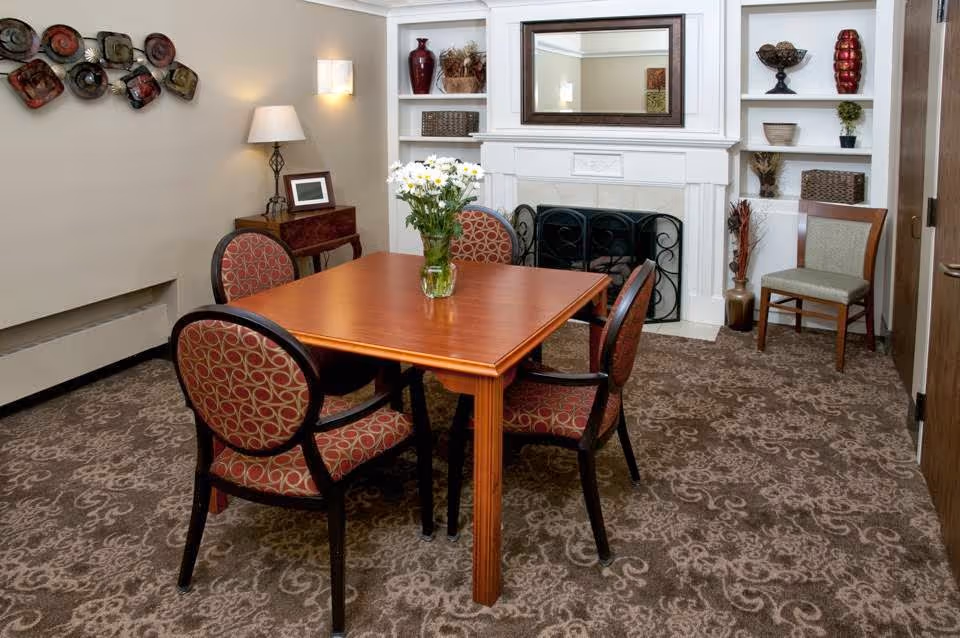 A cozy room with a wooden table surrounded by four upholstered chairs with patterned fabric. A vase with white flowers sits on the table. The room features a white fireplace with a mirror above it and built-in shelves decorated with vases and baskets. There is a patterned carpet on the floor and a small side table with a lamp and picture frame against the wall.
