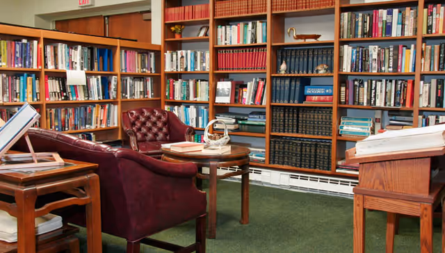 A cozy library room with wooden bookshelves filled with books lining the walls. There are two burgundy leather armchairs and a round wooden table in the center with a basket and some books on it. Additional wooden tables with books and documents are also visible. The floor is carpeted in green.