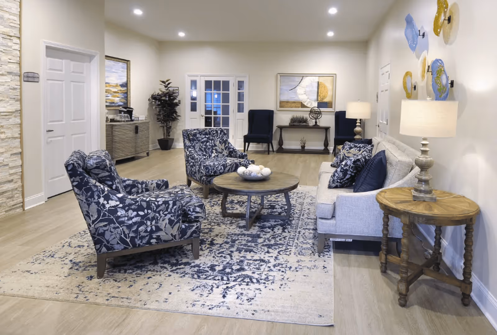 A well-lit living room with a beige sofa adorned with dark blue and patterned cushions, two matching patterned armchairs, a round wooden coffee table with decorative balls, and a patterned area rug. The room features wooden flooring, a side table with a lamp, wall art, and two dark blue chairs against the far wall near a set of French doors.