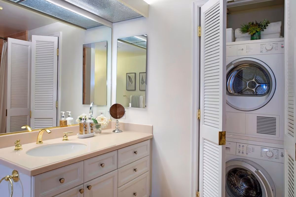 Bathroom vanity with a sink, gold faucet, soap dispensers, a small mirror, and flowers on the countertop. To the right, there is a closet with louvered doors containing a stacked washer and dryer. A large mirror is mounted above the vanity, reflecting part of the bathroom and a towel rack with framed artwork on the wall.