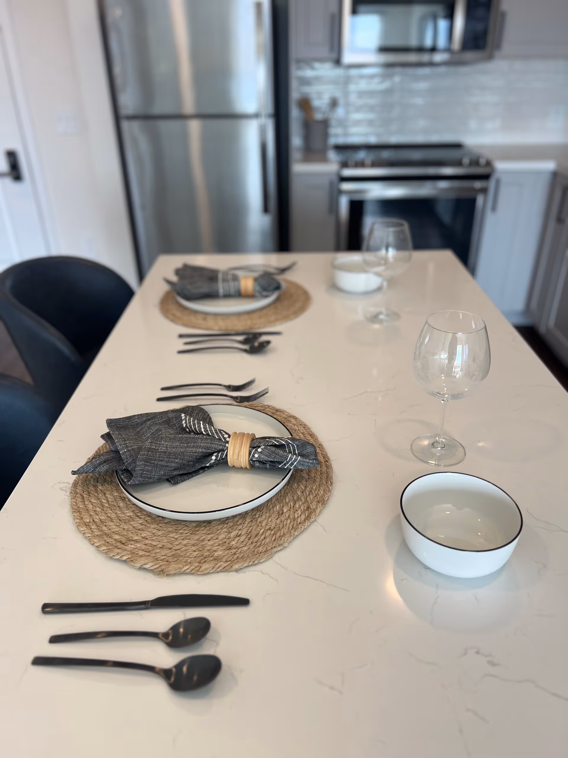 A modern kitchen island set for two with white plates, dark cloth napkins with wooden rings, black cutlery, wine glasses, and a white bowl. In the background, there is a stainless steel refrigerator, stove, and microwave with white cabinetry and a tiled backsplash.