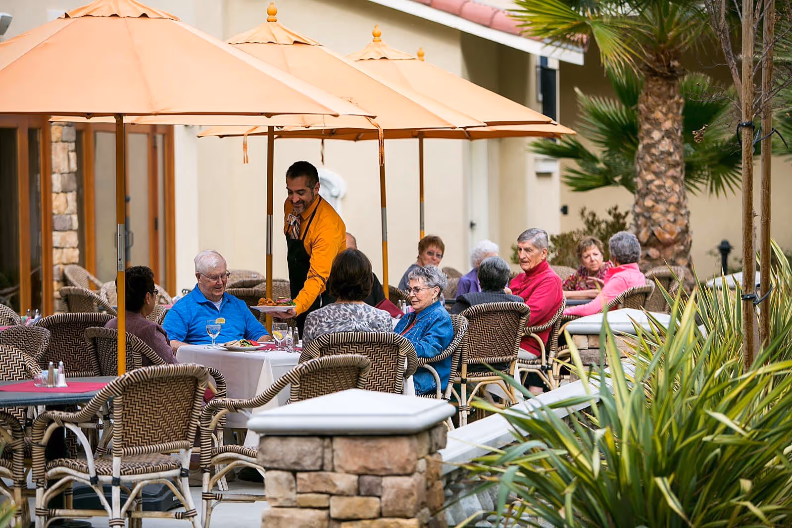 A group of elderly people sitting at outdoor tables under large beige umbrellas, enjoying a meal served by a waiter in a yellow shirt and black apron. The setting is a patio area with wicker chairs, stone pillars, and palm trees, suggesting a warm and pleasant environment.