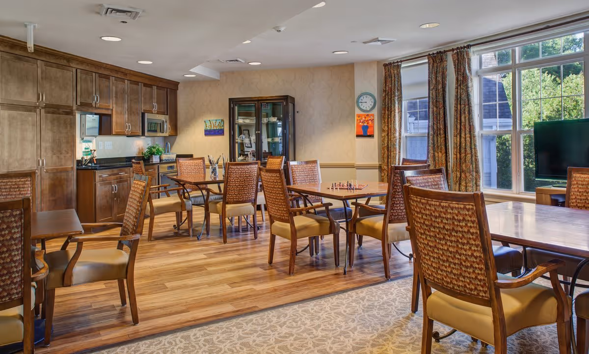 Bright communal dining/activity room with wooden tables and chairs, a kitchenette along the left wall, and large windows letting in daylight.