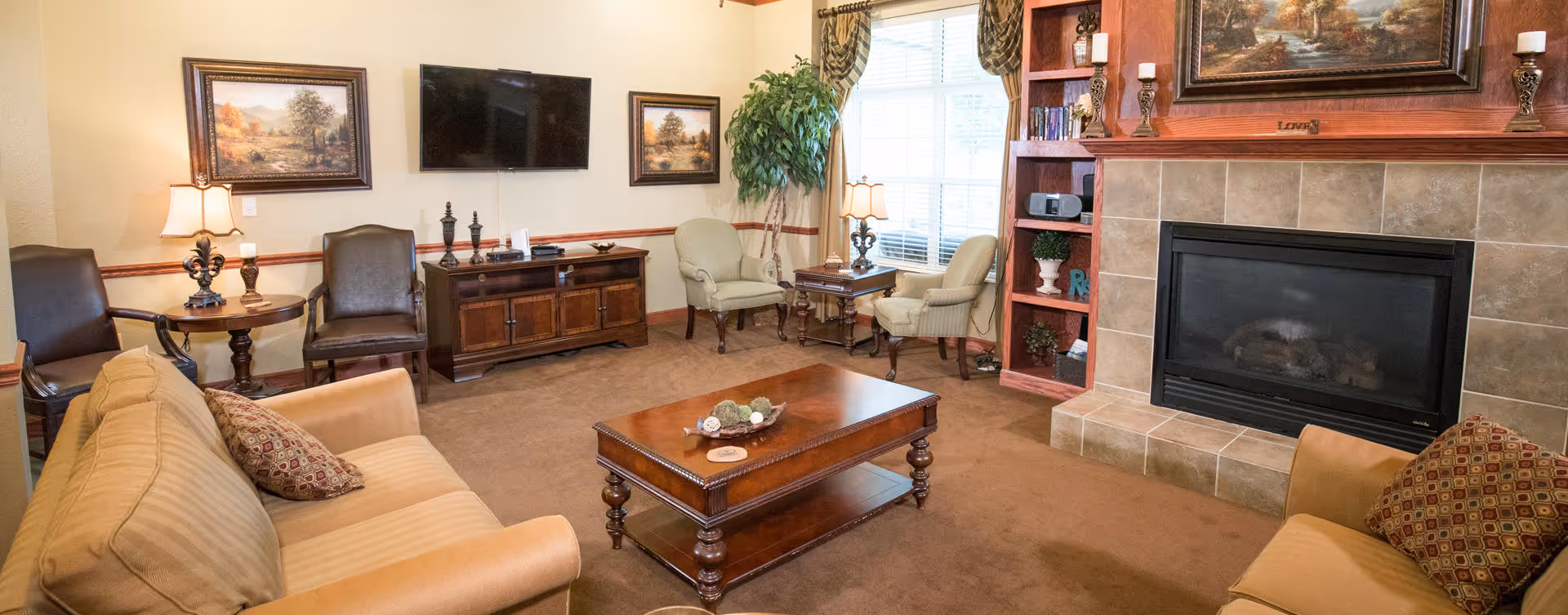 A cozy living room with beige and brown tones featuring a beige sofa and armchairs with patterned cushions, a wooden coffee table, a fireplace with a tiled surround, built-in shelves with decorative items, a flat-screen TV mounted on the wall, framed landscape paintings, and a large window with curtains allowing natural light to fill the room.