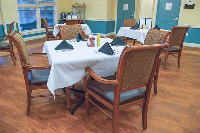 A dining room with wooden floors and several tables covered with white tablecloths. Each table is set with black folded napkins, silverware, and condiments including ketchup and mustard. The room has large windows with white blinds and teal-colored lower walls with yellow upper walls.