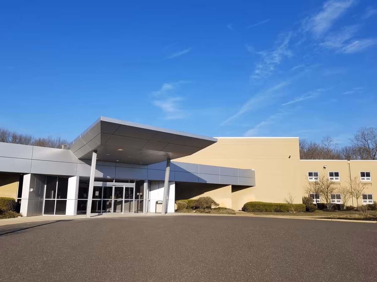 Exterior front view of a modern building with a covered entrance, large glass doors, beige walls, and a clear blue sky above. There are some bushes and small trees around the building.