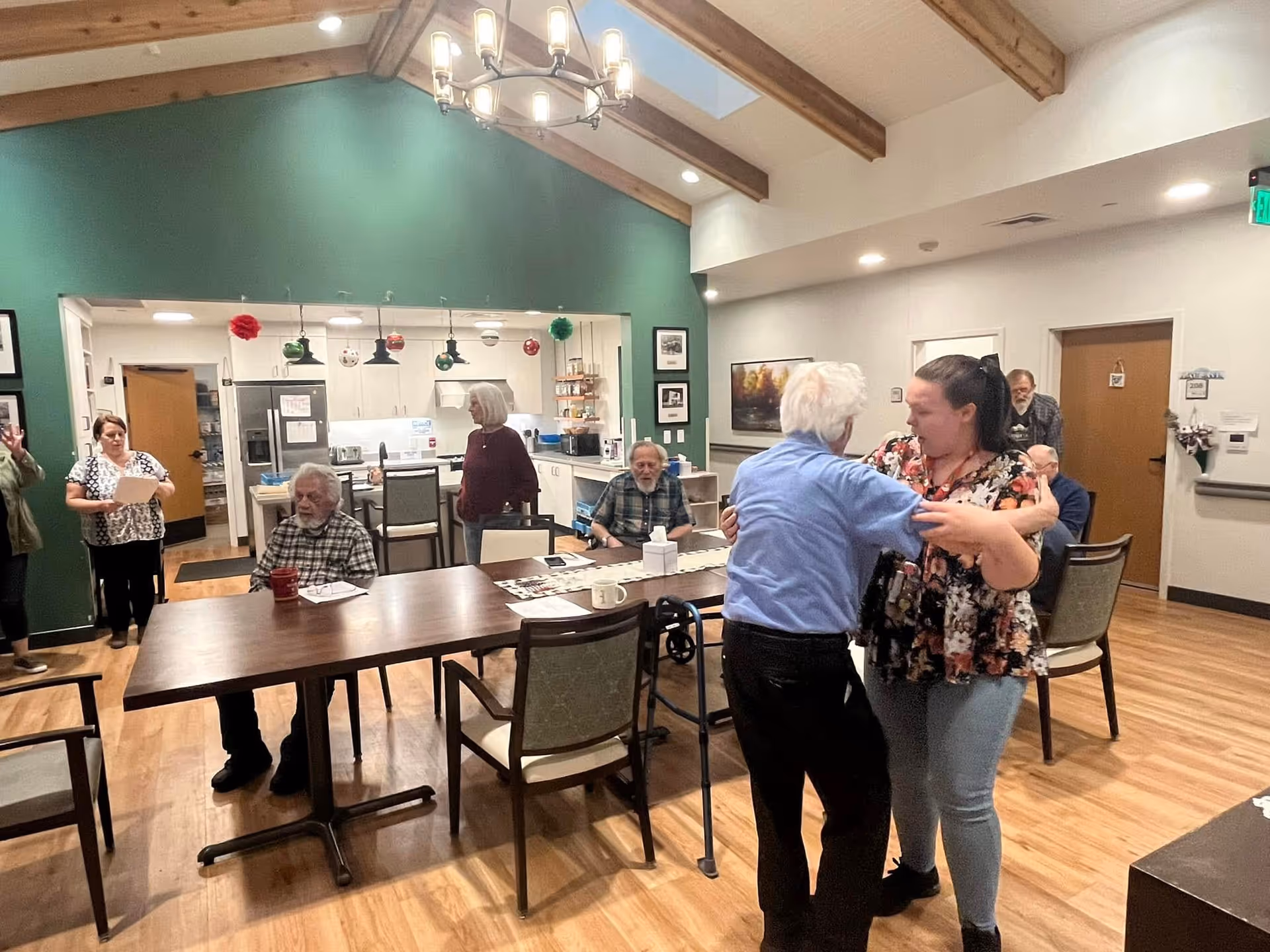 A group of elderly people and a caregiver are in a communal room with wooden floors and a green accent wall. Two people are dancing near the center, while others are seated or standing around a large wooden table. The room has a high ceiling with exposed wooden beams and a chandelier. The kitchen area is visible in the background.