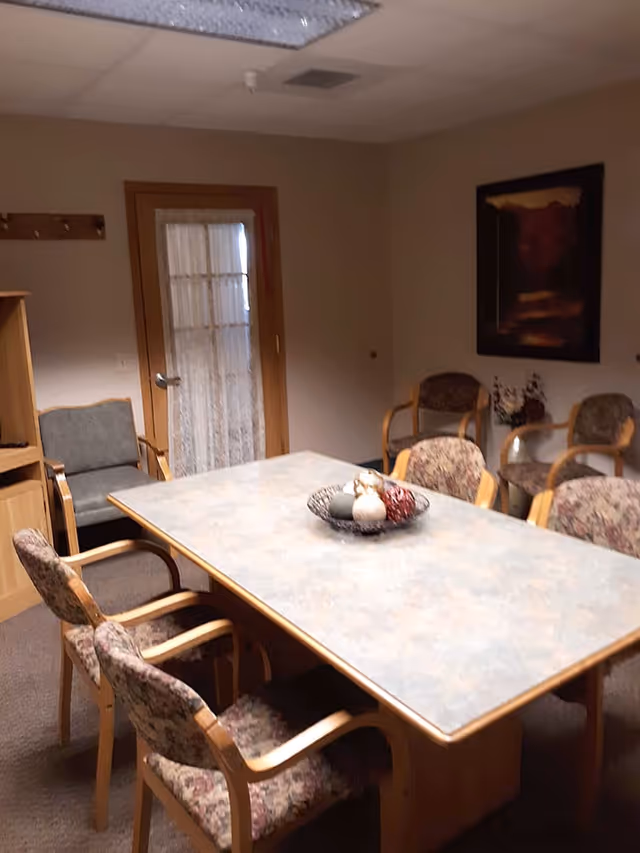 A small meeting or dining room with a rectangular table surrounded by six floral-patterned chairs. The room has beige walls, a wooden door with a lace curtain, a wooden cabinet, and a framed painting on the wall. A decorative bowl with ornamental items is placed on the table.