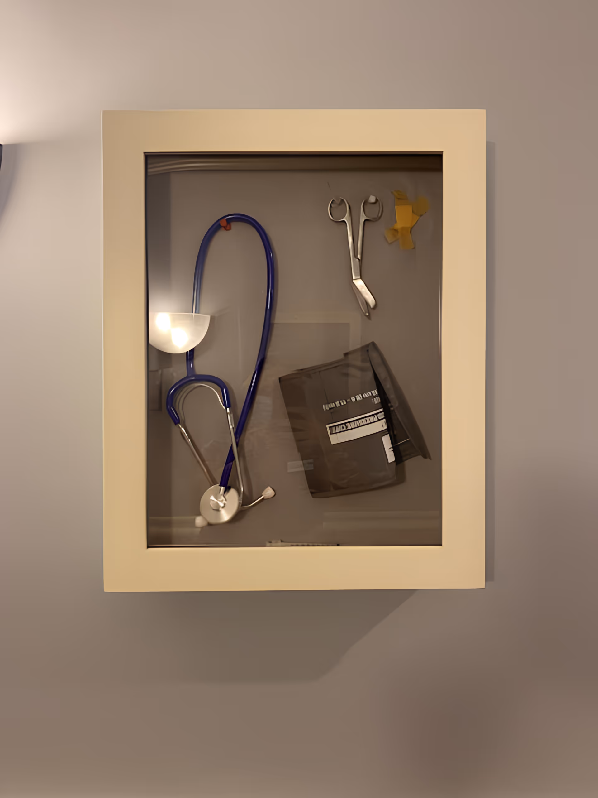 A beige wall-mounted medical cabinet with a glass front containing a blue stethoscope, a pair of medical scissors, some yellow adhesive tape, and a blood pressure cuff.