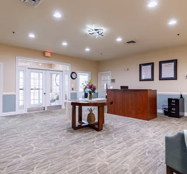 Reception area of a senior living facility with a wooden front desk, a round table with a flower vase and decorative pineapple, beige walls, carpeted floor, and glass doors leading outside.