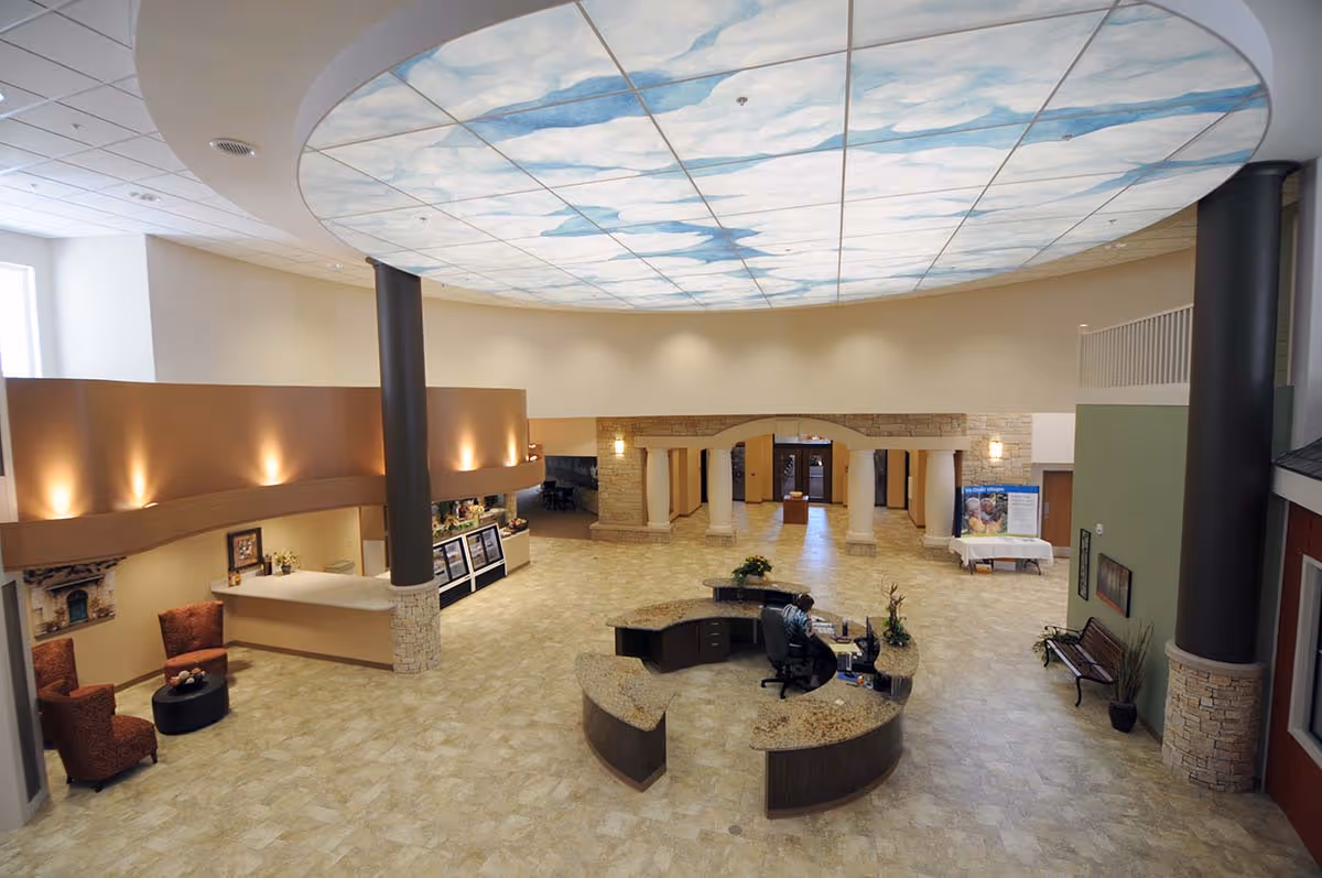 Spacious senior living lobby with a circular reception desk, seating areas, stone columns and a large sky-themed ceiling panel.