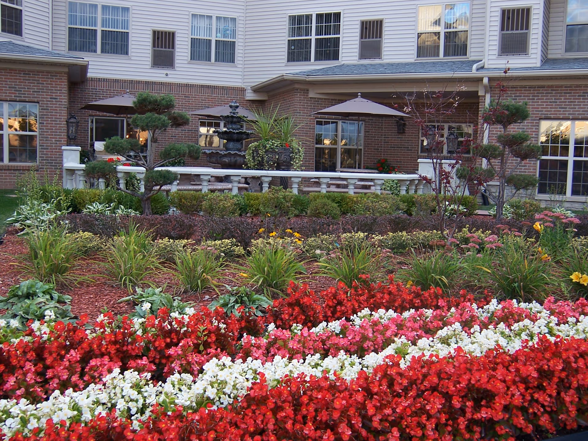 A vibrant garden with rows of red, white, and pink flowers in front of a senior living facility building. The building has brick and siding exterior with multiple windows. There are two patio umbrellas and a decorative fountain on a raised patio area surrounded by greenery and small trees.
