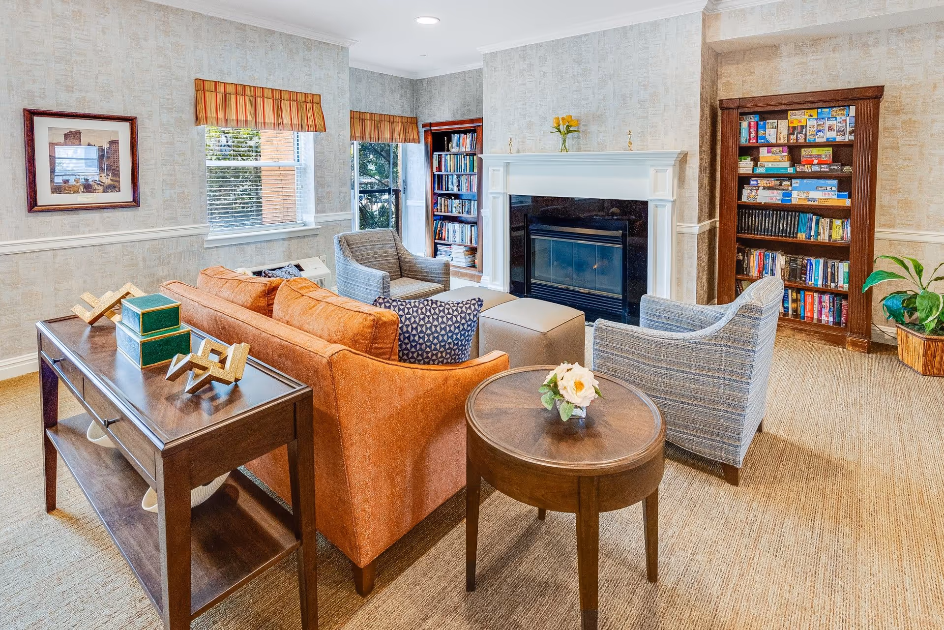 A bright common living room with an orange sofa, armchairs, a fireplace, bookshelves, and side tables.