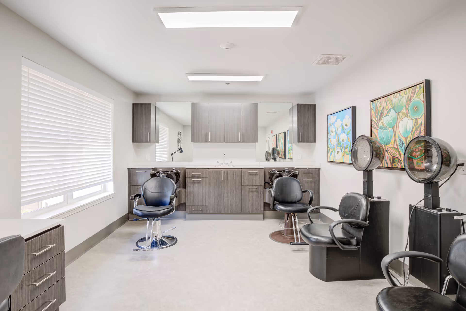 A bright and clean hair salon area with three black salon chairs, two hair dryers, and a counter with cabinets and a sink. The room has white walls, a large window with blinds, and colorful floral paintings on the wall.