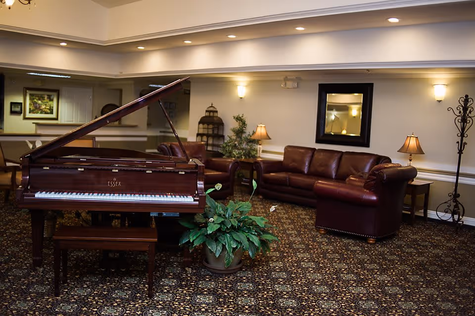 A cozy living room area in an assisted living facility featuring a grand piano with an open lid, a matching piano bench, a large potted plant in front of the piano, and a seating area with a brown leather sofa and armchair. The room is warmly lit with wall sconces and table lamps, and decorated with a framed mirror, a coat rack, and a patterned carpet.