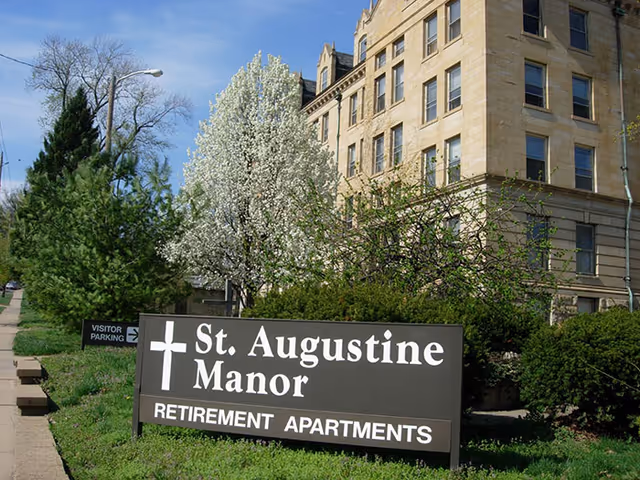 Exterior view of St. Augustine Manor retirement apartments building with a large sign in front displaying the facility name and a cross symbol. The building is multi-story with many windows, surrounded by green bushes and trees, including a tree with white blossoms under a clear blue sky.