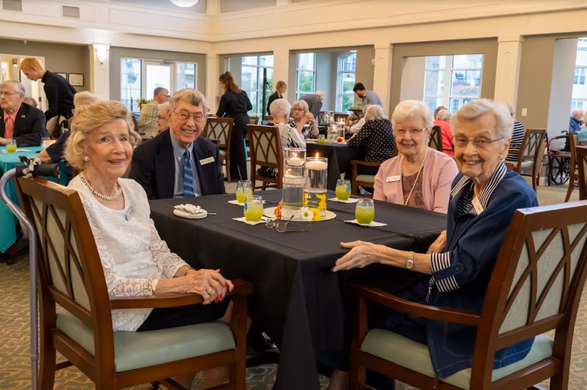 Four elderly individuals sitting around a table with a black tablecloth in a dining room, smiling at the camera. The table has three floating candles in glass holders and four glasses with a green beverage. Other people are seated and standing in the background in a well-lit room with large windows.