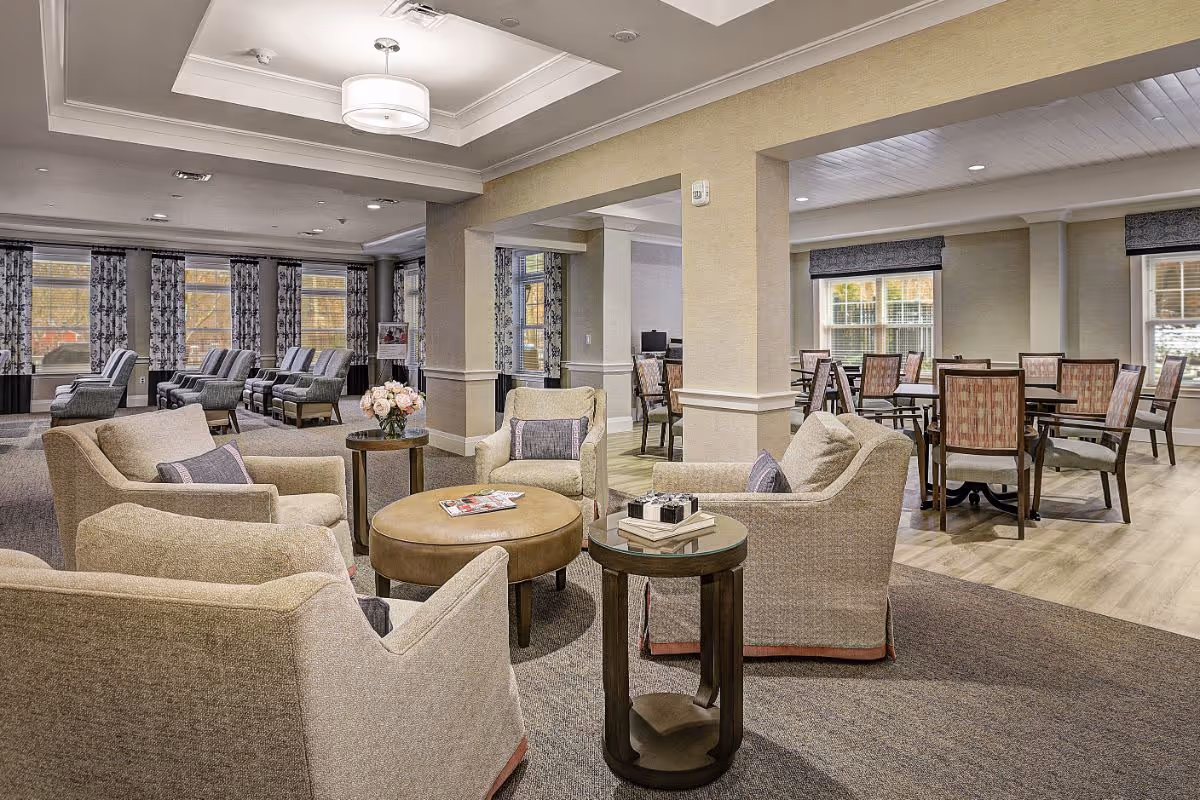 Comfortable senior living common area with upholstered armchairs grouped around coffee tables and a dining area in the background.