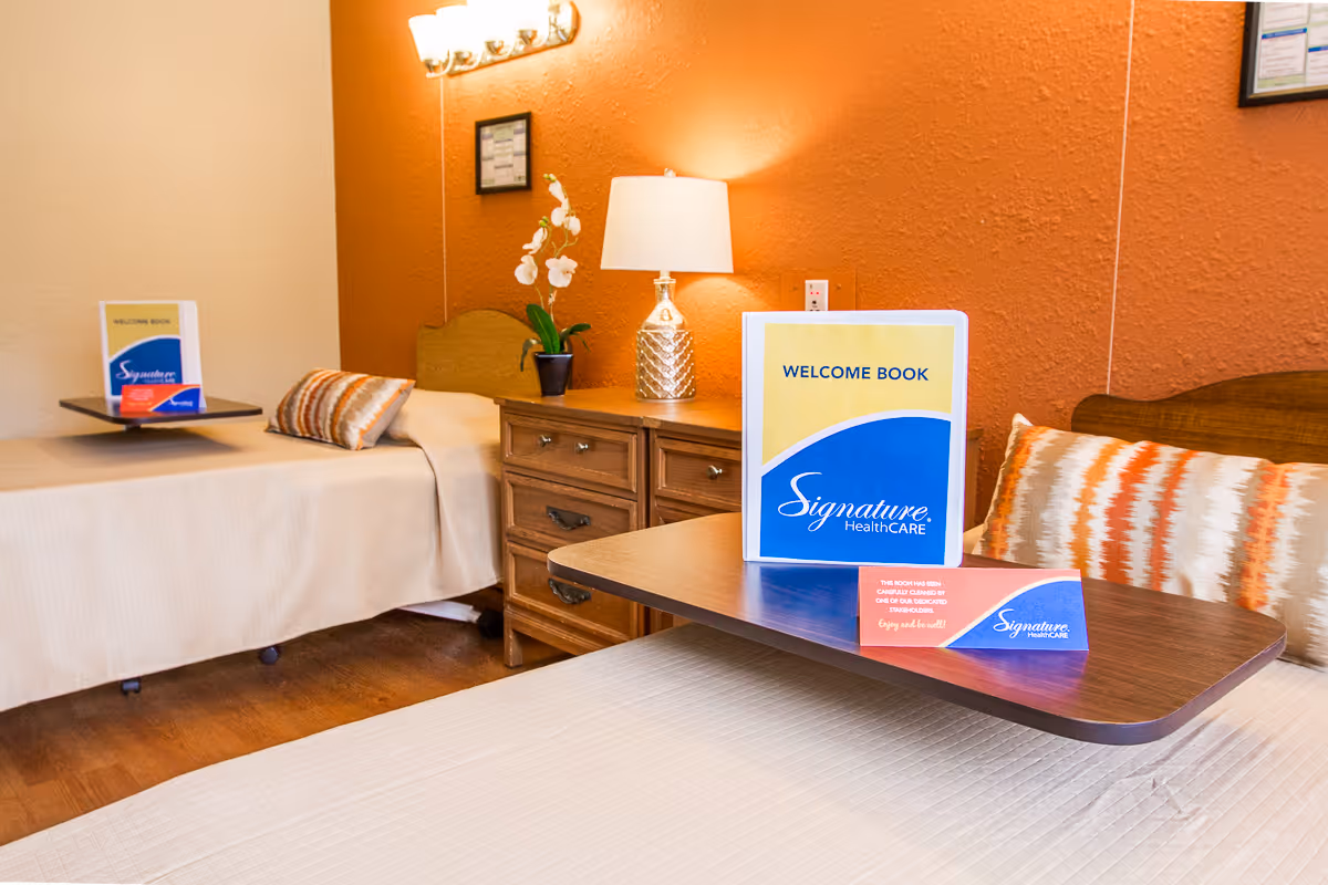 Two beds in a senior living facility room with a nightstand, lamp, decorative pillows, and a 'Welcome Book' displayed on a tray table.