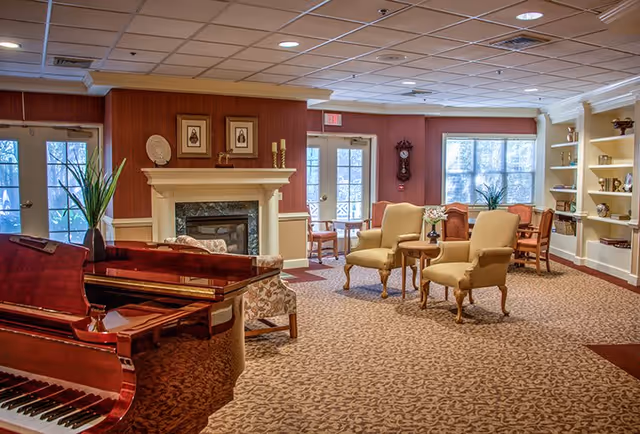 Cozy communal living room with a grand piano, fireplace, upholstered chairs, and tables.