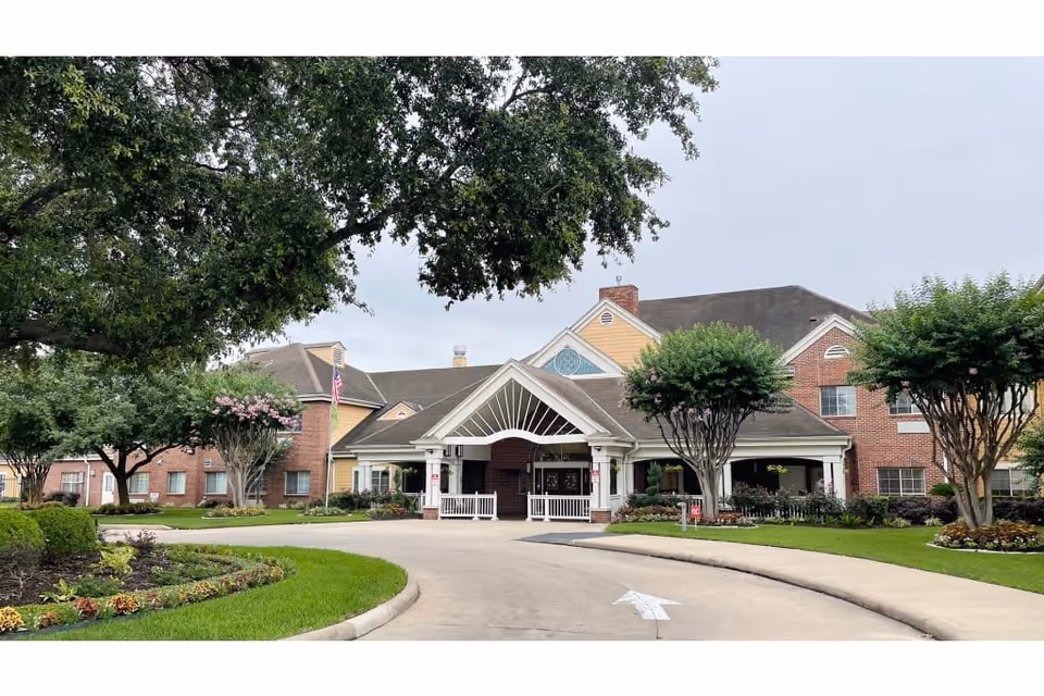 Front exterior view of a senior living facility named Atria Westchase, showing a driveway leading to the main entrance with a covered porch. The building is two stories with a combination of brick and siding, surrounded by well-maintained landscaping including trees and flower beds.