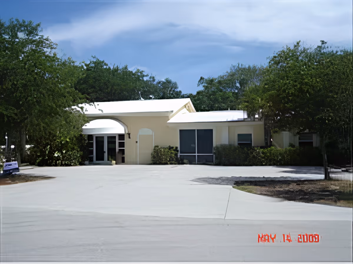 Exterior view of a single-story building with a white roof and light-colored walls, surrounded by trees and bushes. The building has large windows and a covered entrance. The sky is partly cloudy.