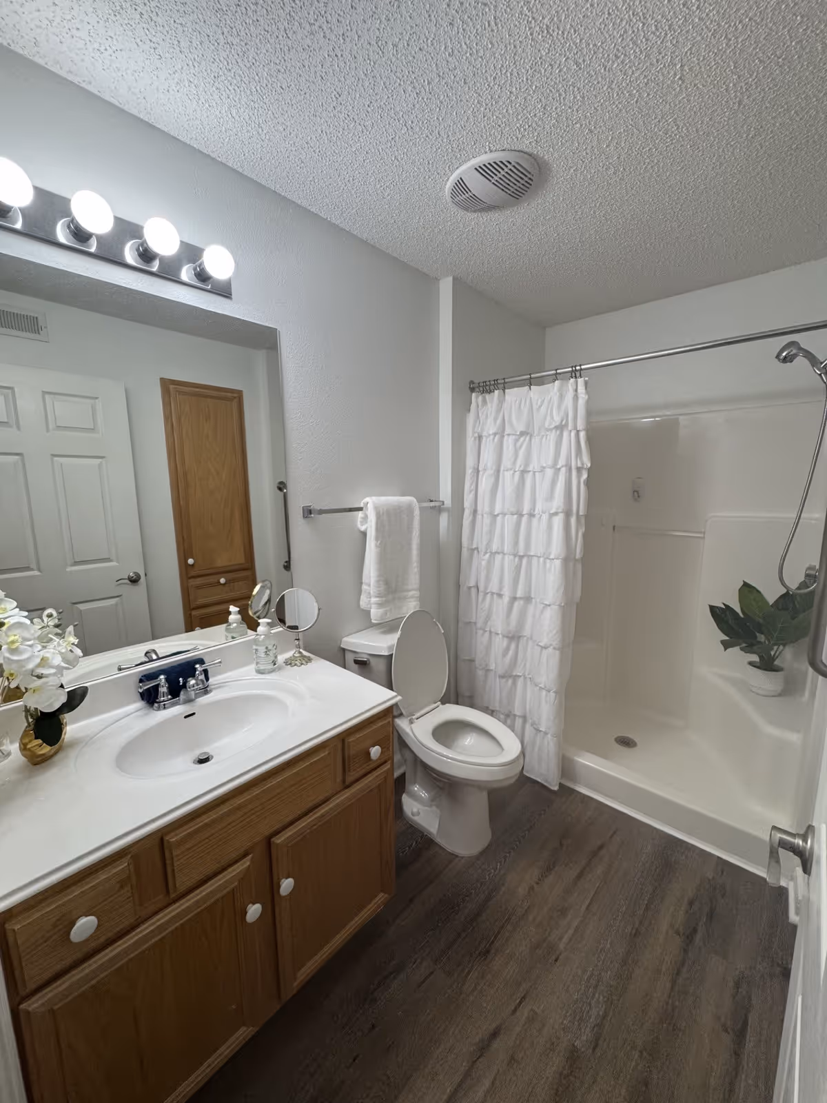 A clean bathroom with a wooden vanity featuring a white sink and countertop. Above the sink is a large mirror with four round lights. To the right of the vanity is a toilet with the lid open, and next to it is a shower with a white ruffled shower curtain. The floor is a dark wood laminate, and there is a small plant placed inside the shower. A towel hangs on a rack above the toilet.