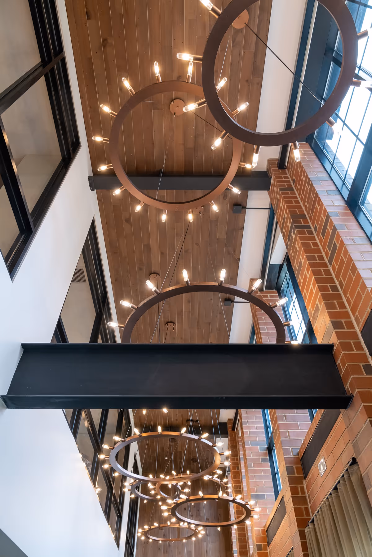 View looking up at a high ceiling with wooden panels, large circular chandeliers with multiple exposed light bulbs, black metal beams, and large windows with black frames and brick walls.