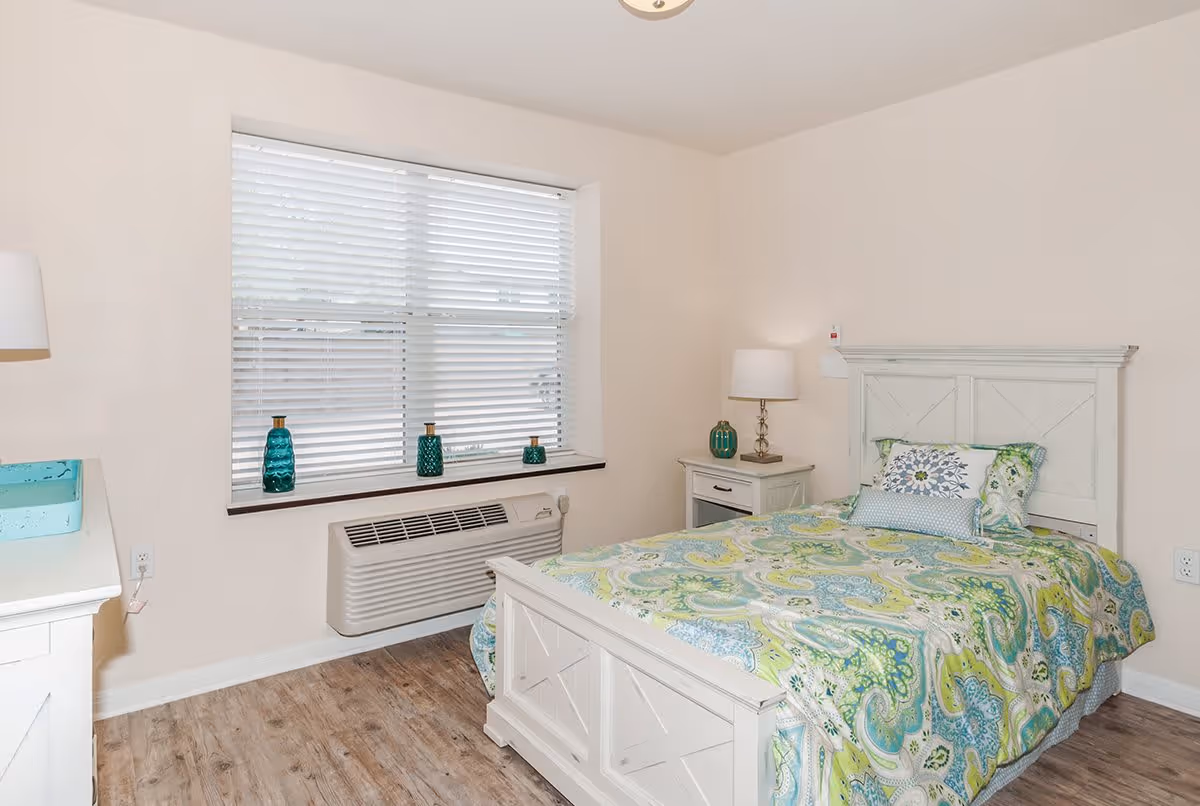 A bright bedroom with a white wooden bed frame and a colorful green and blue patterned bedspread. There is a window with white blinds above a wall-mounted air conditioning unit. A white nightstand with a lamp and decorative items is next to the bed, and a white dresser is partially visible on the left side of the image. The floor is wood-style laminate.