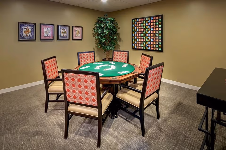 A game room with a round table covered with a green felt surface designed for playing cards, surrounded by six wooden chairs with red patterned upholstery. The room has beige walls decorated with framed artwork and a large colorful alphabet poster. A potted plant is placed in the corner.