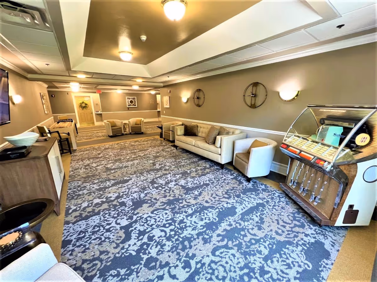 A spacious and well-lit common area with patterned carpet flooring, beige walls, and ceiling lights. The room features a beige sofa with cushions, two beige armchairs, a vintage-style jukebox, and a wooden cabinet with a bowl on top. There are decorative wall clocks and framed artwork on the walls, and a hallway with additional seating and a door with a wreath in the background.