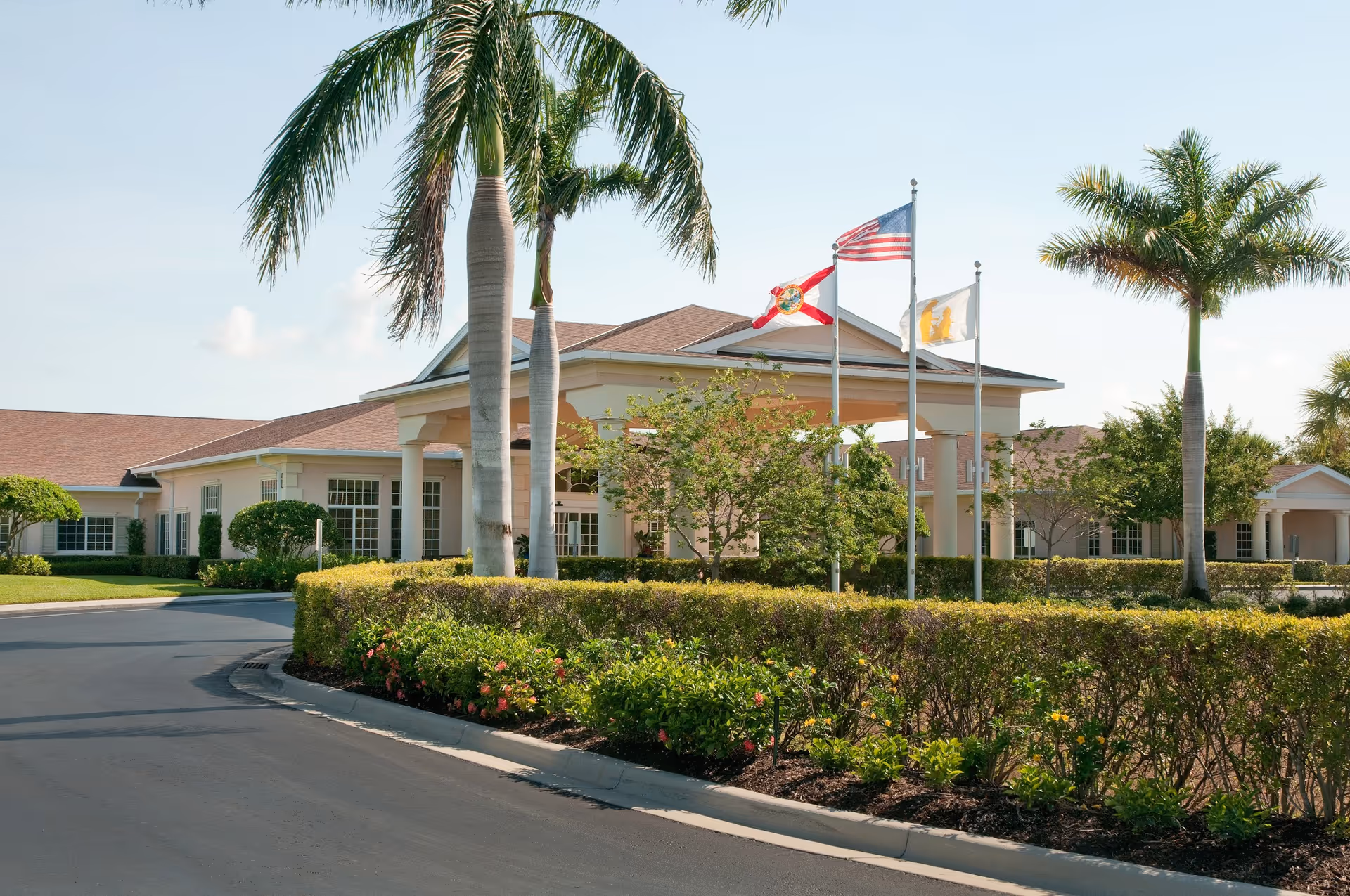 Front entrance of a low-rise care facility with a porte-cochere, three flagpoles, palm trees, and manicured landscaping.