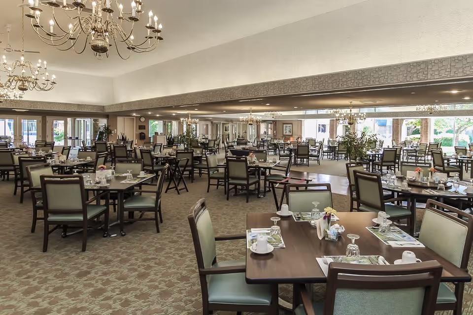A spacious dining room with multiple tables and chairs arranged neatly. Each table is set with placemats, cups, glasses, and small flower vases. The room features large windows allowing natural light to brighten the space, and elegant chandeliers hang from the ceiling.