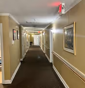 A long indoor hallway in a senior living facility with beige walls, brown carpet, framed artwork on the walls, handrails along both sides, ceiling lights, and exit signs at the far end.