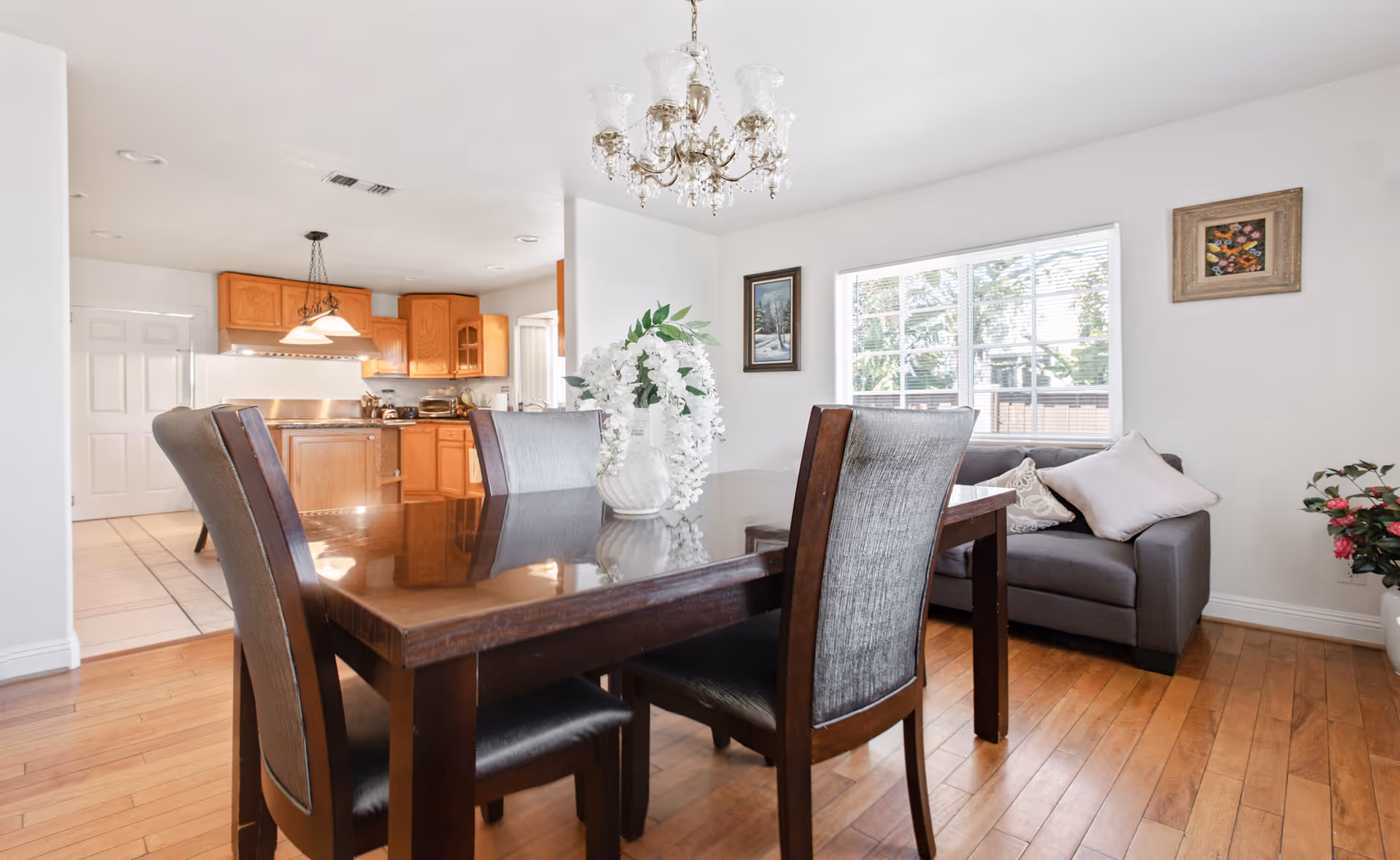 A bright dining area with a dark wooden table and four upholstered chairs. A white vase with white flowers is placed on the table. In the background, there is a kitchen with wooden cabinets and a white door. To the right, there is a gray sofa with cushions near a window with blinds. The room has wooden flooring and white walls with framed pictures. A chandelier hangs above the dining table.