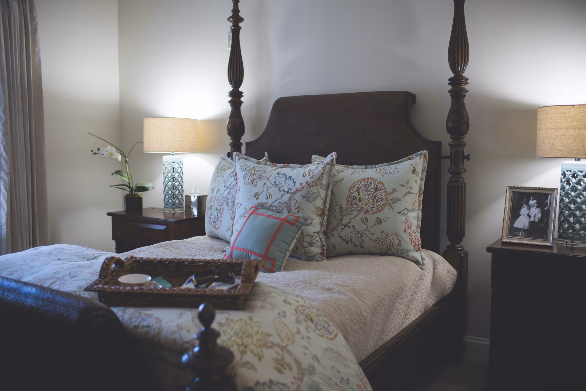 A cozy bedroom with a dark wooden four-poster bed adorned with floral patterned pillows and a light-colored quilt. There are two bedside tables, each with a lamp featuring a textured blue base and beige shade. One bedside table has a small plant and a glass of water, while the other has a framed black and white photograph. A woven tray with a cup and some items rests on the bed.
