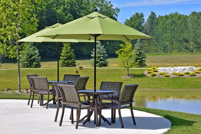 Outdoor patio area with two black metal tables and eight matching chairs under two large green umbrellas. The patio overlooks a small pond with grassy banks, trees, and a clear blue sky in the background.