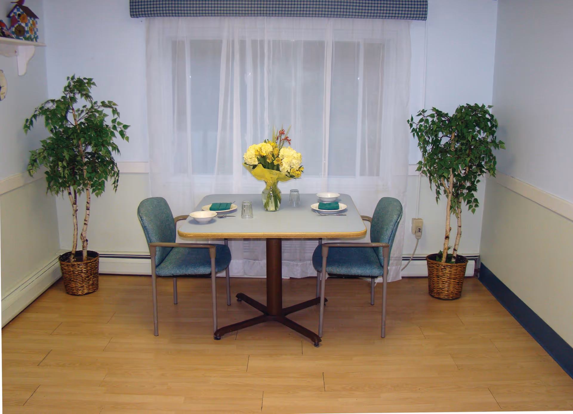 A small dining area with a square table set for two, featuring two blue upholstered chairs. The table has a vase with yellow flowers, two bowls, two plates with green napkins, and two upside-down glasses. The room has light-colored walls, a window with sheer white curtains and a blue valance, and two potted plants on either side of the table. The floor is light wood.