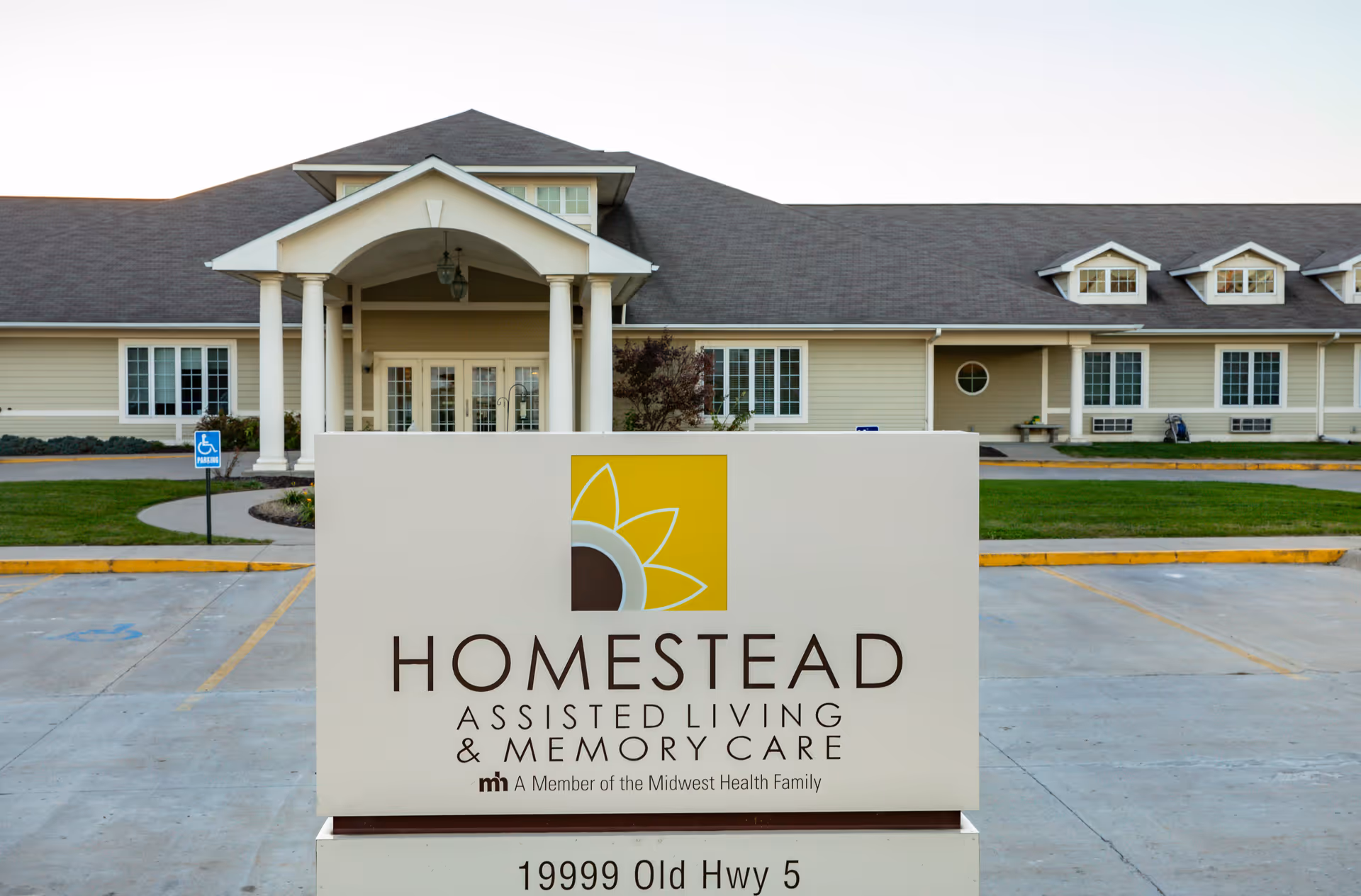 Exterior view of Homestead Assisted Living & Memory Care of Centerville building with a large sign in the foreground displaying the facility's name and address. The building has a beige exterior with white trim, multiple windows, and a covered entrance supported by columns. There is a parking lot with marked spaces and a handicap parking sign visible.